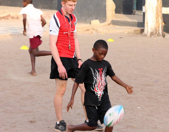 A child kicks a rugby ball as his coach watches on